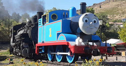A colorful cartoon train with a smiling face, alongside a classic steam locomotive, set in a scenic outdoor area.