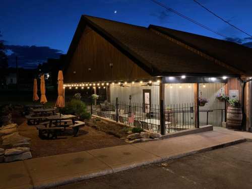 A cozy restaurant exterior at night, featuring outdoor seating, string lights, and orange umbrellas under a twilight sky.