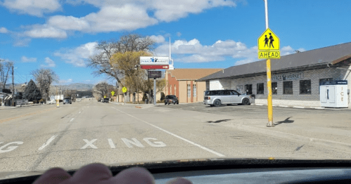 A view of a quiet street with a market, a crosswalk sign, and a clear blue sky with scattered clouds.