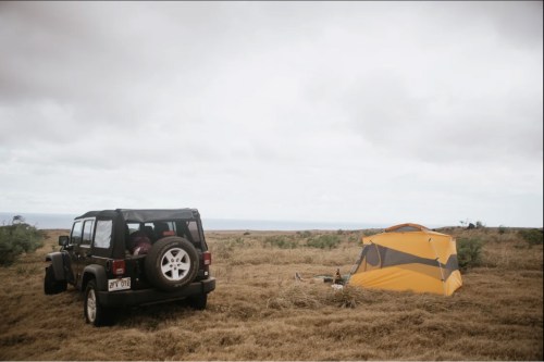 A black SUV parked beside a yellow tent in a grassy area with a cloudy sky and ocean in the background.