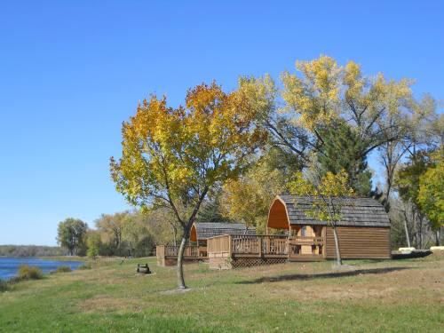 Two wooden cabins by a calm lake, surrounded by trees with autumn foliage under a clear blue sky.