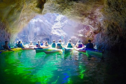 A group of kayakers paddles through a cave, surrounded by shimmering green water and illuminated by natural light.