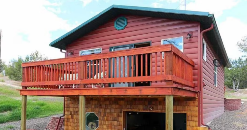 A rustic red cabin with a wooden deck, surrounded by greenery and gravel, featuring a garage underneath.