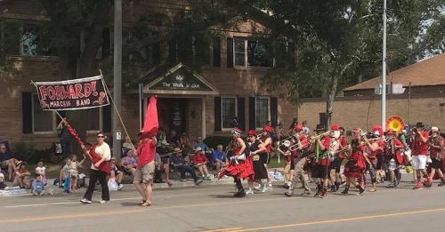 A marching band in colorful outfits parades down the street, with spectators watching from the sidelines.