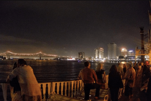 Nighttime view of a waterfront with people enjoying the scenery and a brightly lit city skyline in the background.