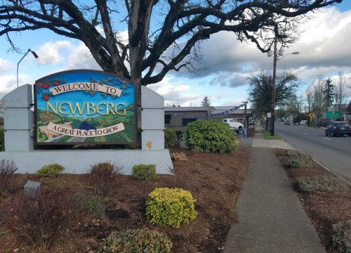 Welcome sign for Newberg, featuring colorful text and landscaping along a sidewalk beside a road.
