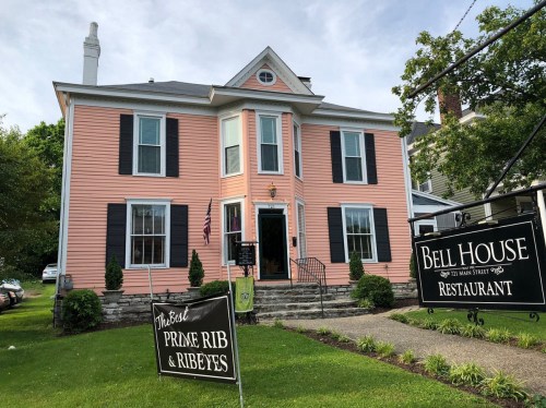 A pink house with black shutters, featuring signs for "Bell House Restaurant" and "The Best Prime Rib & Ribeyes."