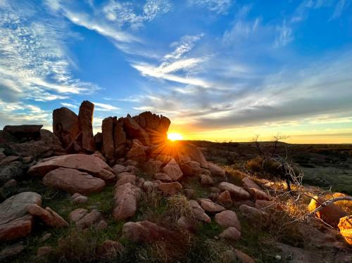 Sunset behind rocky formations, casting warm light over a grassy landscape under a vibrant sky.