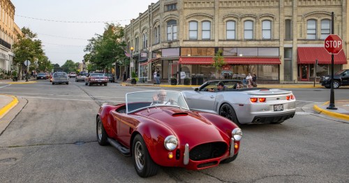 Two classic cars, a red convertible and a silver sports car, drive through a charming downtown street with historic buildings.