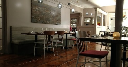 A cozy restaurant interior with wooden floors, tables set for dining, and soft lighting from hanging fixtures.