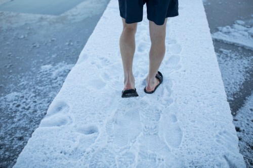 A person walks barefoot on a salt-covered path near water, leaving footprints in the salt.