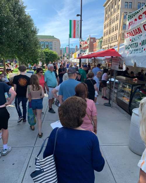 A bustling street fair with people browsing food stalls under sunny skies and colorful banners.