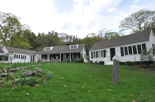 A white, two-story house with a porch, surrounded by green grass and trees, featuring a smaller adjacent building.