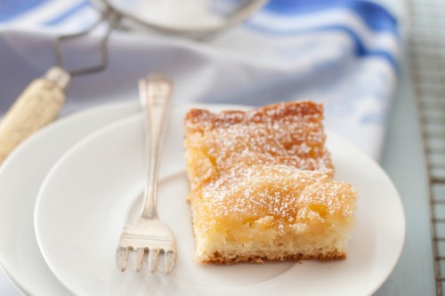 A slice of cake dusted with powdered sugar on a white plate, with a fork beside it. Soft blue cloth in the background.