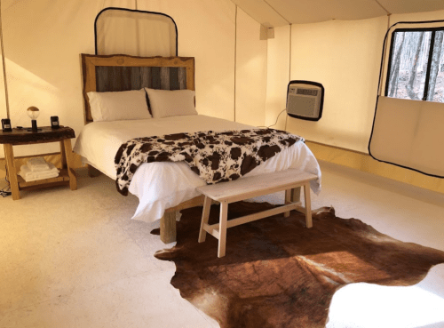 Cozy glamping tent interior with a bed, wooden furniture, and a cowhide rug on the floor. Natural light from windows.