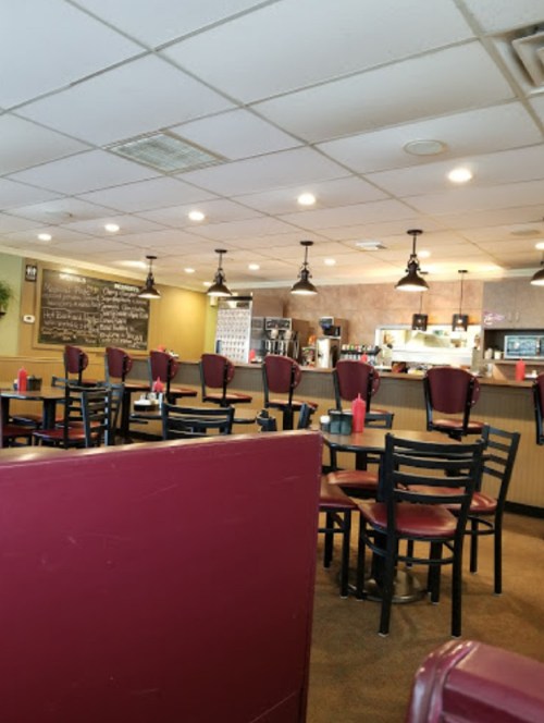 A cozy diner interior with empty tables, red chairs, and a counter displaying food and drinks. Warm lighting throughout.