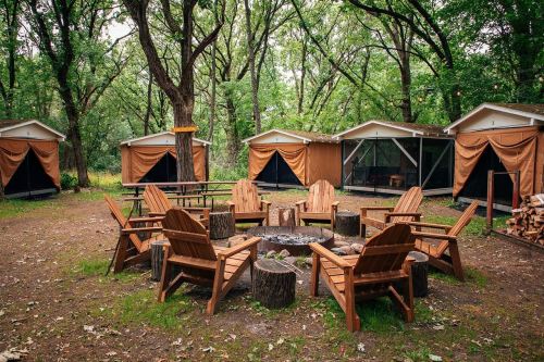 A campsite with wooden chairs around a fire pit, surrounded by tents and lush green trees.