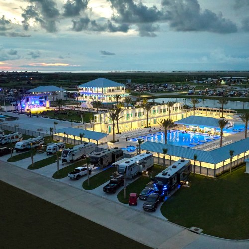 Aerial view of a resort with a pool, palm trees, RVs, and a stage, set against a sunset sky.