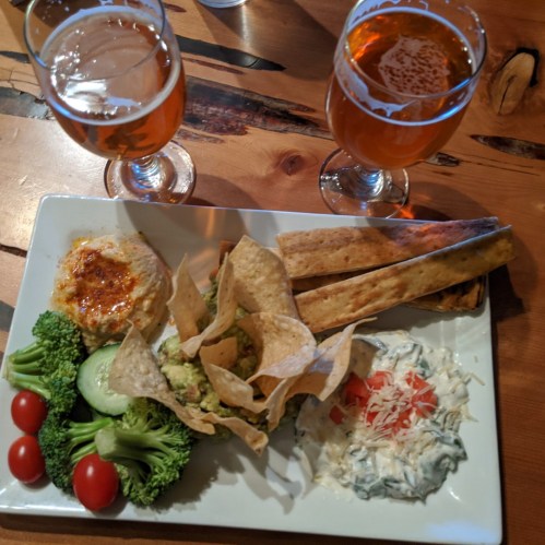 A platter with guacamole, hummus, chips, veggies, and a dip, accompanied by two glasses of amber beer.