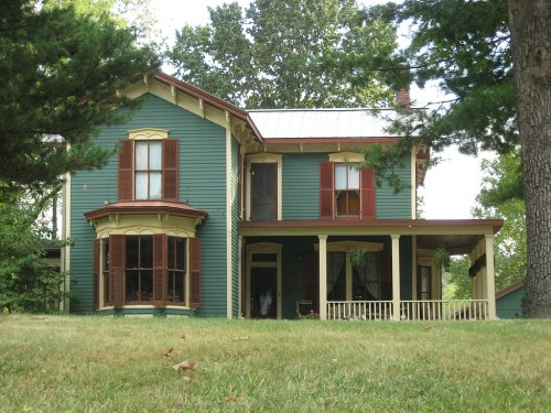 A two-story Victorian-style house with green siding, red shutters, and a porch, surrounded by trees and grass.