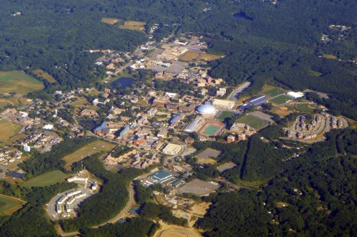 Aerial view of a small town with buildings, sports fields, and green areas surrounded by trees and fields.