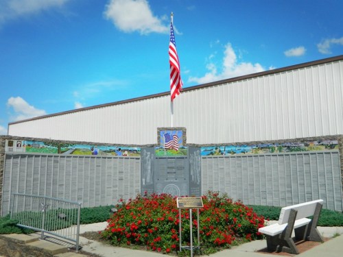 A memorial with an American flag, surrounded by flowers and a stone wall featuring engraved names and images.