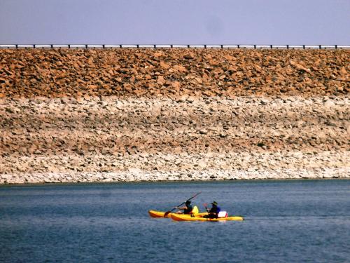 Two people kayaking on a calm body of water, with a rocky dam in the background under a clear sky.