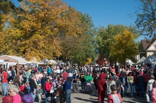 A bustling outdoor market scene with a large crowd, colorful autumn trees, and vendor tents under a clear blue sky.