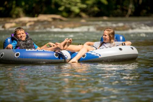 Two children relax on inflatable tubes in a river, enjoying a sunny day surrounded by greenery.