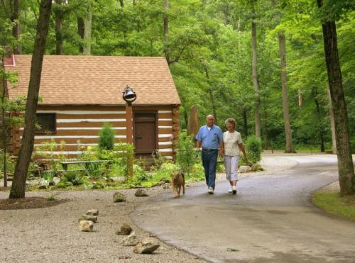 A couple walks a dog along a path near a log cabin in a lush, green forest.