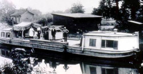 Black and white photo of a houseboat docked by a riverside, with people and buildings in the background.