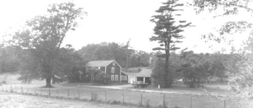 A black and white image of a farmhouse surrounded by trees and a fence in a rural setting.