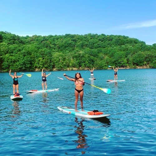 A group of women paddleboarding on a calm lake surrounded by green trees under a clear blue sky.