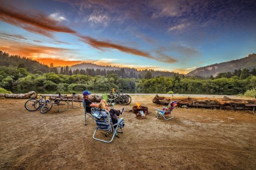A group of people relaxes by a campfire near a lake, surrounded by trees and mountains at sunset. Bikes are nearby.