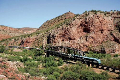 A train crosses a bridge in a rugged landscape of red rock formations and green vegetation.