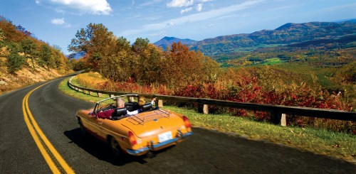 A convertible car drives along a winding road with vibrant autumn foliage and mountains in the background.