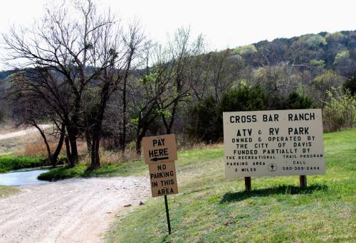 Sign for Cross Bar Ranch ATV & RV Park, with a "Pay Here" sign and a no parking notice, surrounded by trees and a dirt road.
