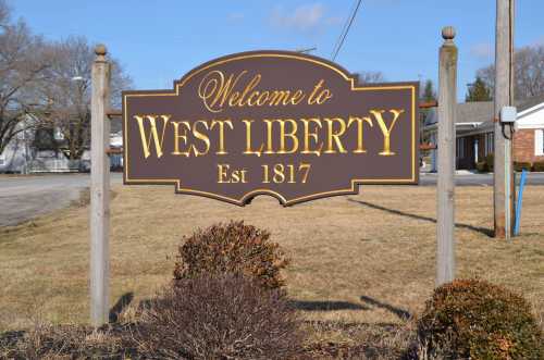 Sign welcoming visitors to West Liberty, established in 1817, with a grassy area and buildings in the background.