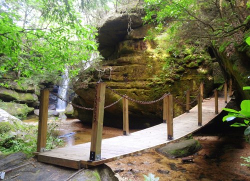 A wooden bridge with a chain railing crosses a serene stream surrounded by lush green foliage and rocky cliffs.