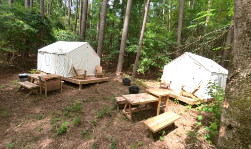 Two white tents in a wooded area, surrounded by trees, with wooden tables and chairs set up nearby.