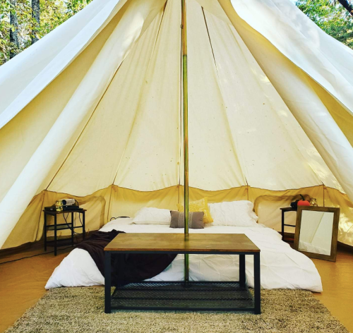 Cozy glamping tent interior with a bed, wooden table, and decorative items, surrounded by nature.