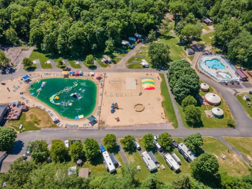 Aerial view of a recreational area with a pool, sandy beach, playground, and camping trailers surrounded by trees.