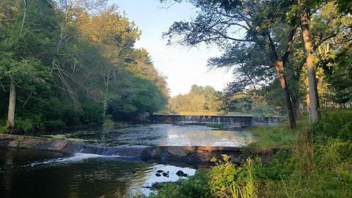 A serene landscape featuring a calm river, a small waterfall, and lush greenery under a clear blue sky.
