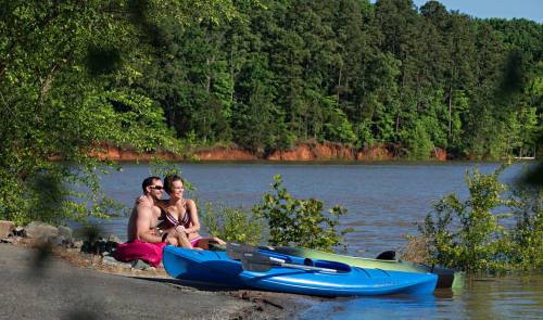 Two people sit by a lake, enjoying the view, with a kayak nearby and lush greenery in the background.