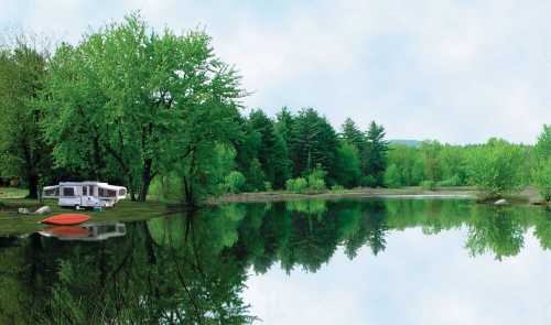 A serene lakeside scene with a camper, lush green trees, and calm water reflecting the landscape.