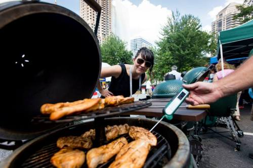 A woman in sunglasses inspects grilled chicken on a barbecue, with a thermometer in hand and city buildings in the background.