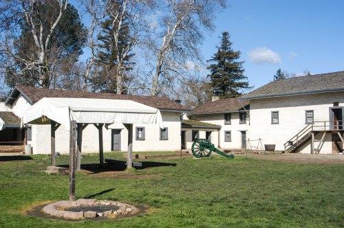 Historic buildings with a grassy area, a covered well, and a cannon in a sunny outdoor setting.
