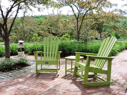 Two green Adirondack chairs and a small table on a brick patio, surrounded by trees and greenery.