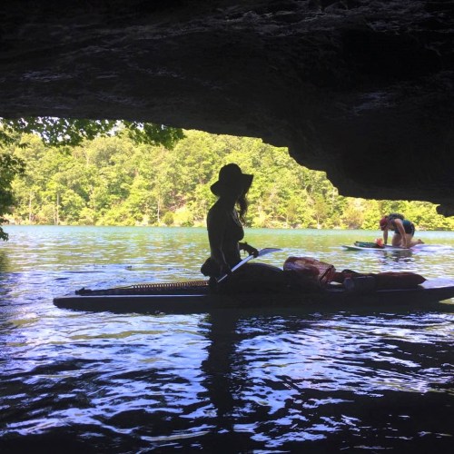 A person in a kayak paddles under a rocky overhang, surrounded by lush greenery and calm water.