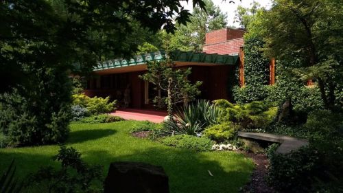 A modern home surrounded by lush greenery and landscaped gardens, featuring a distinctive green roof and brick walls.